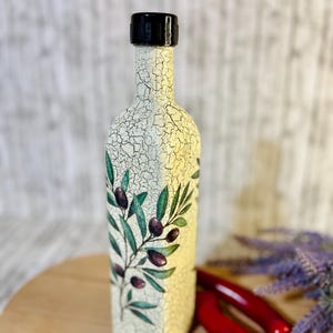 Puede incluir: Una botella de cer&aacute;mica blanca con tapa negra y un dise&ntilde;o pintado de ramas de olivo y aceitunas. La botella est&aacute; sobre una superficie de madera.