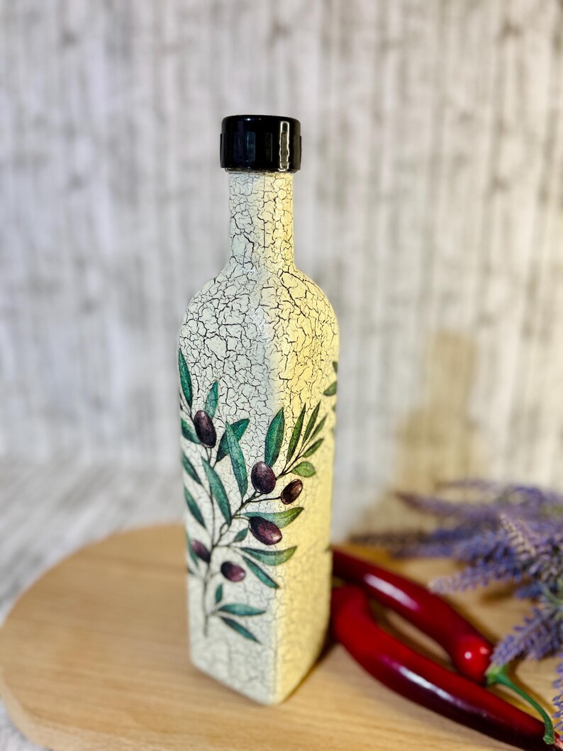 Puede incluir: Una botella de cer&aacute;mica blanca con tapa negra y un dise&ntilde;o pintado de ramas de olivo y aceitunas. La botella est&aacute; sobre una superficie de madera.
