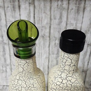 May include: Two white ceramic bottles with a decorative crackle paint finish. One bottle has a green pour spout and the other has a black cap.