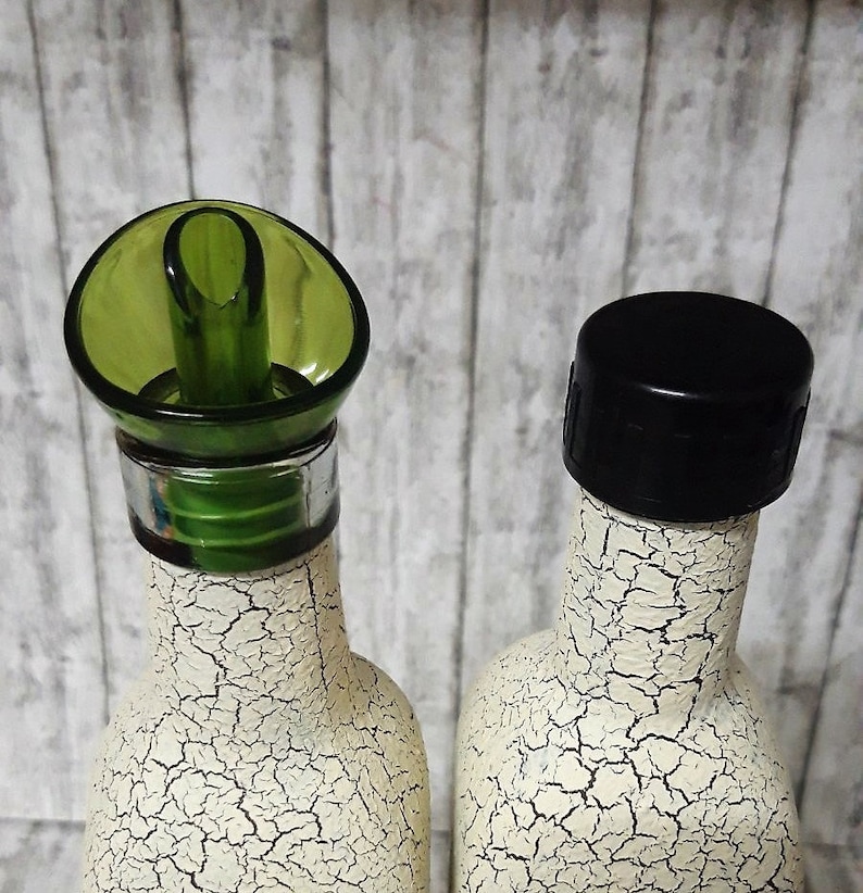 May include: Two white ceramic bottles with a decorative crackle paint finish. One bottle has a green pour spout and the other has a black cap.