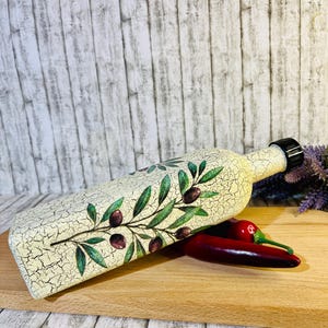 Puede incluir: Una botella de cer&aacute;mica blanca con un acabado desgastado y un dise&ntilde;o de rama de olivo verde. La botella tiene un tap&oacute;n de corcho negro y est&aacute; sobre una tabla de cortar de madera con dos chiles rojos.