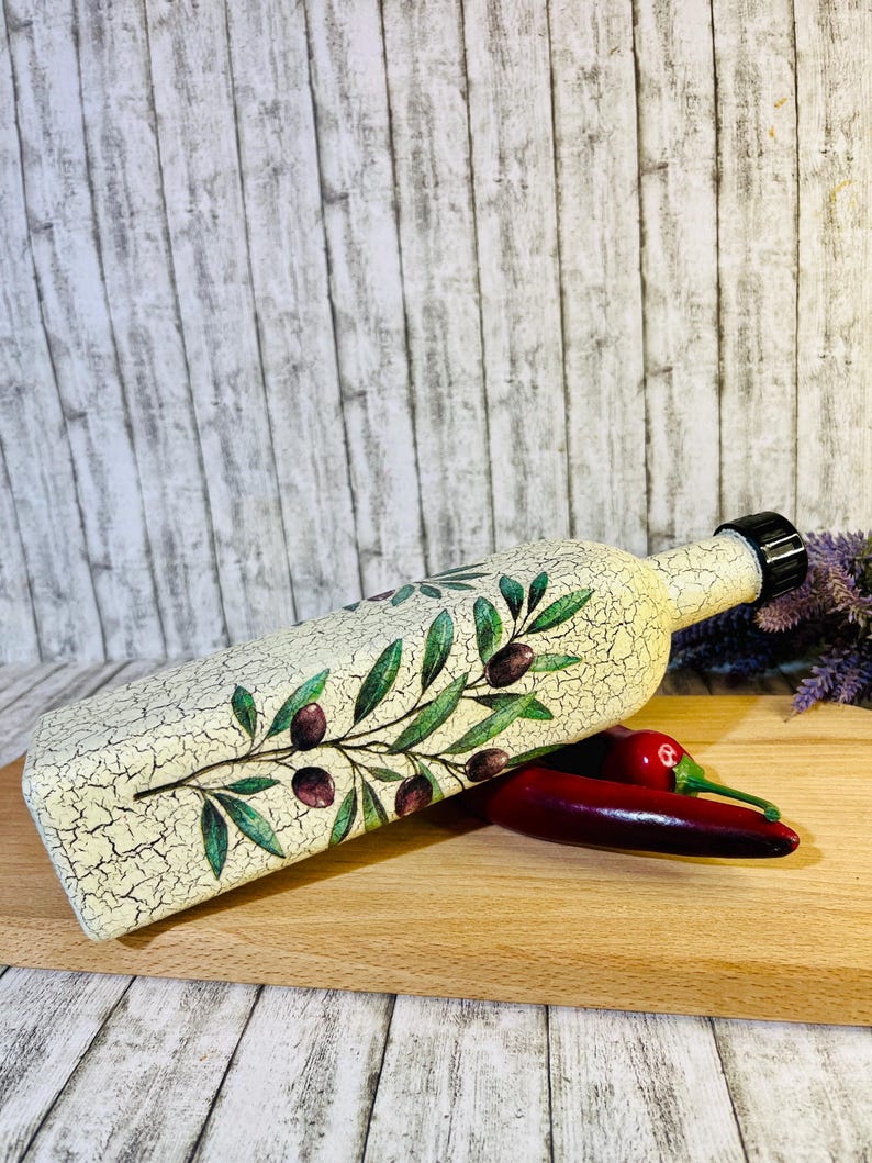 Puede incluir: Una botella de cer&aacute;mica blanca con un acabado desgastado y un dise&ntilde;o de rama de olivo verde. La botella tiene un tap&oacute;n de corcho negro y est&aacute; sobre una tabla de cortar de madera con dos chiles rojos.