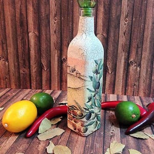 May include: A decorative glass bottle with a green pour spout, featuring a painted design of olive branches and a Tuscan landscape. The bottle is sitting on a wooden surface with red chilli peppers, limes, and bay leaves.