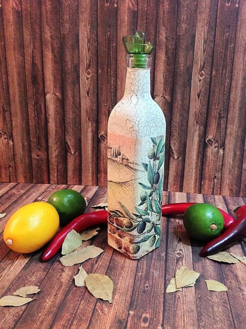 May include: A decorative glass bottle with a green pour spout, featuring a painted design of olive branches and a Tuscan landscape. The bottle is sitting on a wooden surface with red chilli peppers, limes, and bay leaves.