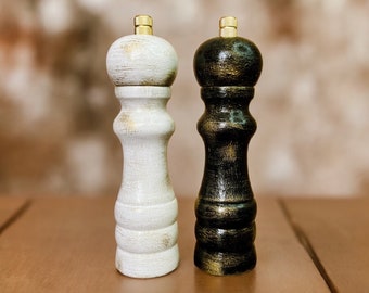 May include: Two wooden salt and pepper mills with a distressed finish. One mill is painted white with gold highlights, and the other is painted black with gold highlights. Both feature a rounded top and a small metallic gold knob at the peak.