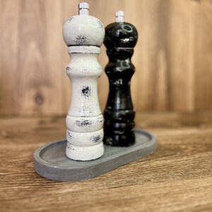 May include: A pair of salt and pepper grinders, one white and one black, both featuring a distressed finish with visible wood grain texture. Each grinder is topped with a silver-colored metal knob and sits on a gray oval tray.