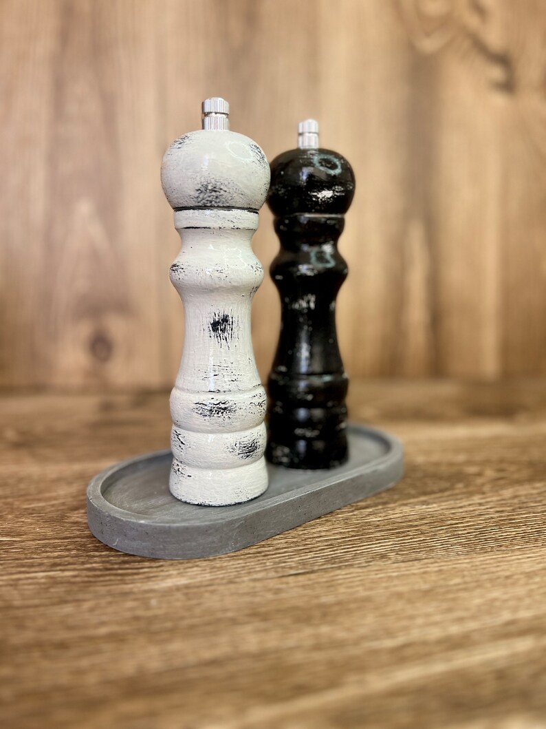 May include: A pair of salt and pepper grinders, one white and one black, both featuring a distressed finish with visible wood grain texture. Each grinder is topped with a silver-colored metal knob and sits on a gray oval tray.