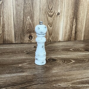 May include: A white pepper mill with a distressed finish and a silver-colored metal knob at the top. The mill features a classic hourglass-shaped body with visible dark wood grain showing through the worn white paint on its surface.