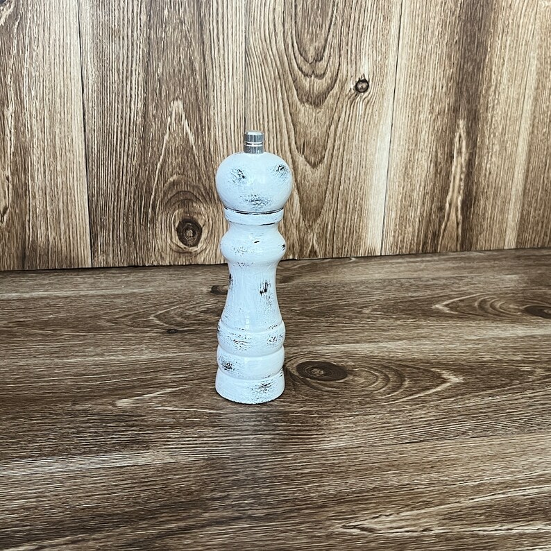 May include: A white pepper mill with a distressed finish and a silver-colored metal knob at the top. The mill features a classic hourglass-shaped body with visible dark wood grain showing through the worn white paint on its surface.
