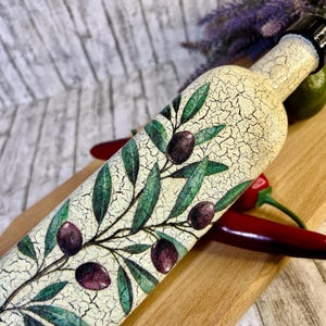 Puede incluir: Una botella de cer&aacute;mica blanca con un acabado de esmalte craquelado, decorada con una rama de olivo verde y aceitunas moradas. La botella tiene una tapa negra y est&aacute; sobre una superficie de madera.