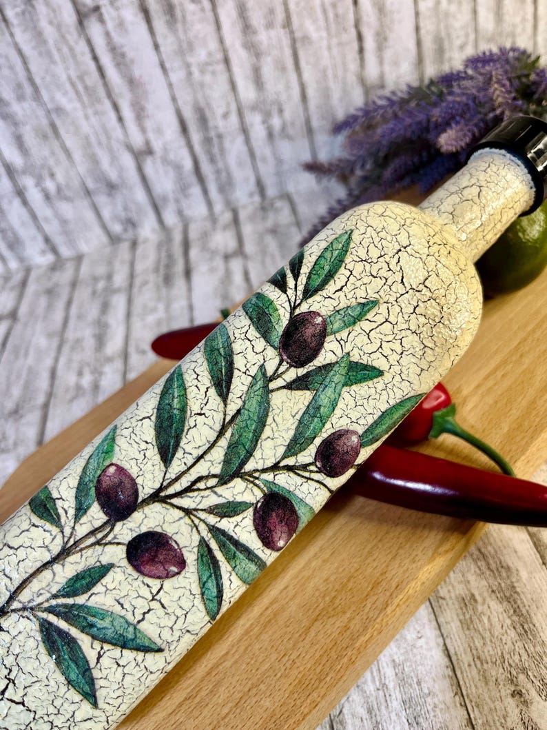 Puede incluir: Una botella de cer&aacute;mica blanca con un acabado de esmalte craquelado, decorada con una rama de olivo verde y aceitunas moradas. La botella tiene una tapa negra y est&aacute; sobre una superficie de madera.