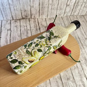 May include: A decorative glass bottle with a floral and avocado design. The bottle has a distressed white finish and a black lid. The bottle is sitting on a wooden cutting board with red chili peppers.
