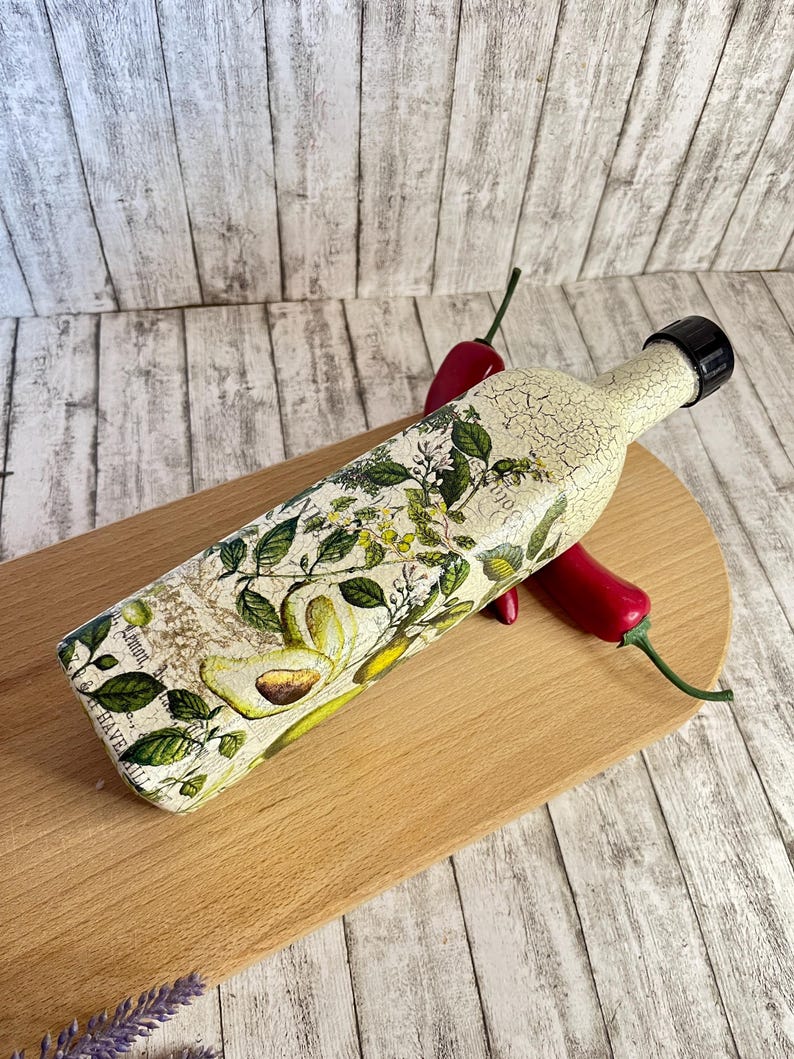 May include: A decorative glass bottle with a floral and avocado design. The bottle has a distressed white finish and a black lid. The bottle is sitting on a wooden cutting board with red chili peppers.