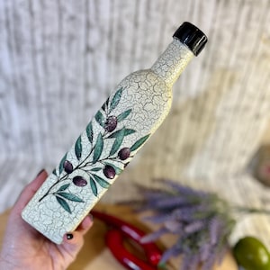 Puede incluir: Una botella de cer&aacute;mica blanca con una tapa negra y un dise&ntilde;o pintado de ramas de olivo con aceitunas moradas. La botella est&aacute; siendo sostenida por una mano.