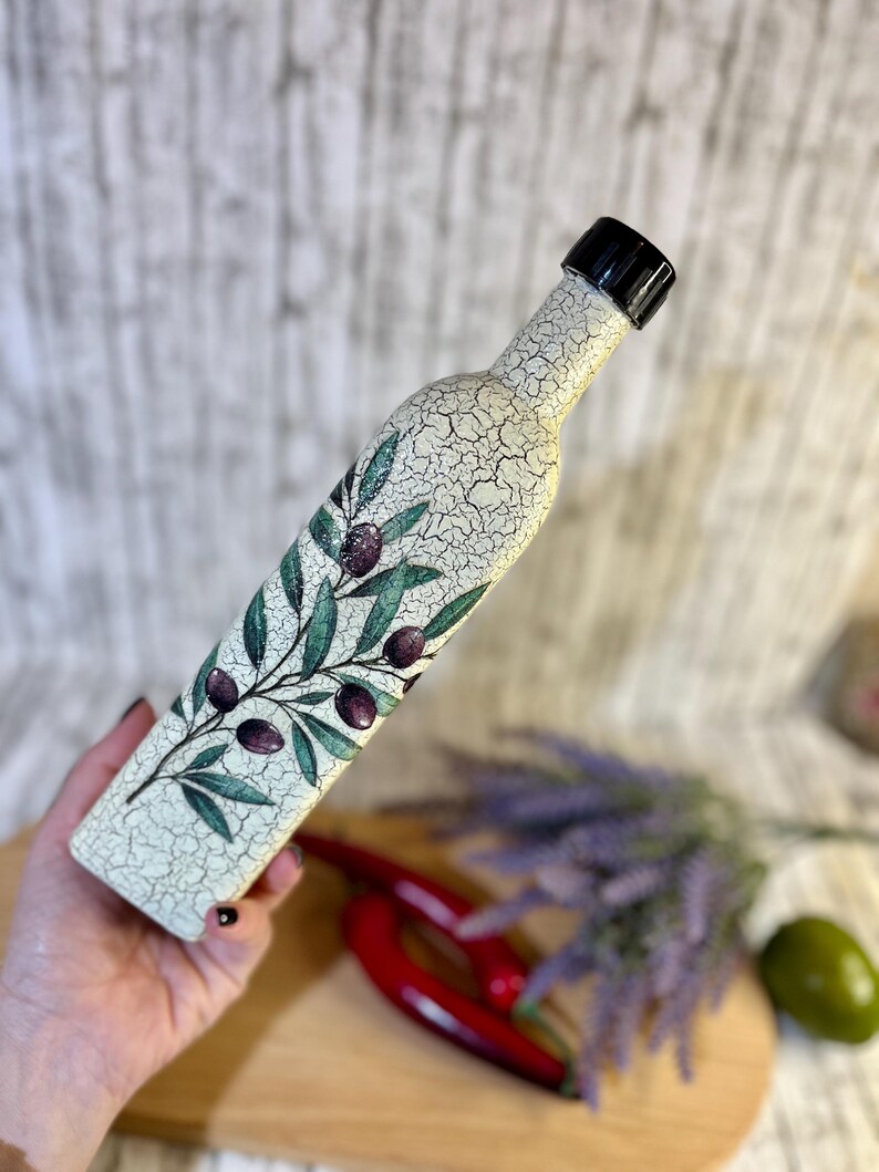 Puede incluir: Una botella de cer&aacute;mica blanca con una tapa negra y un dise&ntilde;o pintado de ramas de olivo con aceitunas moradas. La botella est&aacute; siendo sostenida por una mano.