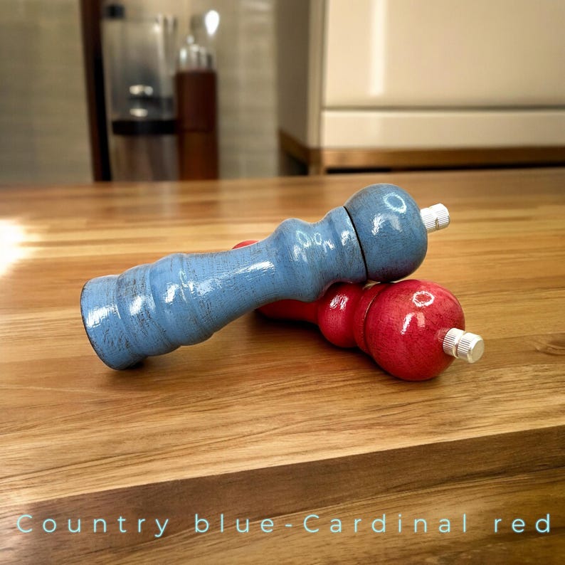 May include: Two wooden salt and pepper shakers, one blue and one red, on a wooden table. The blue shaker is labeled "Country Blue" and the red shaker is labeled "Cardinal Red".