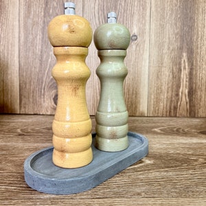 May include: A set of two wooden salt and pepper shakers with a distressed finish. The shakers are in a light yellow and a light green color. The shakers are on a gray oval tray.