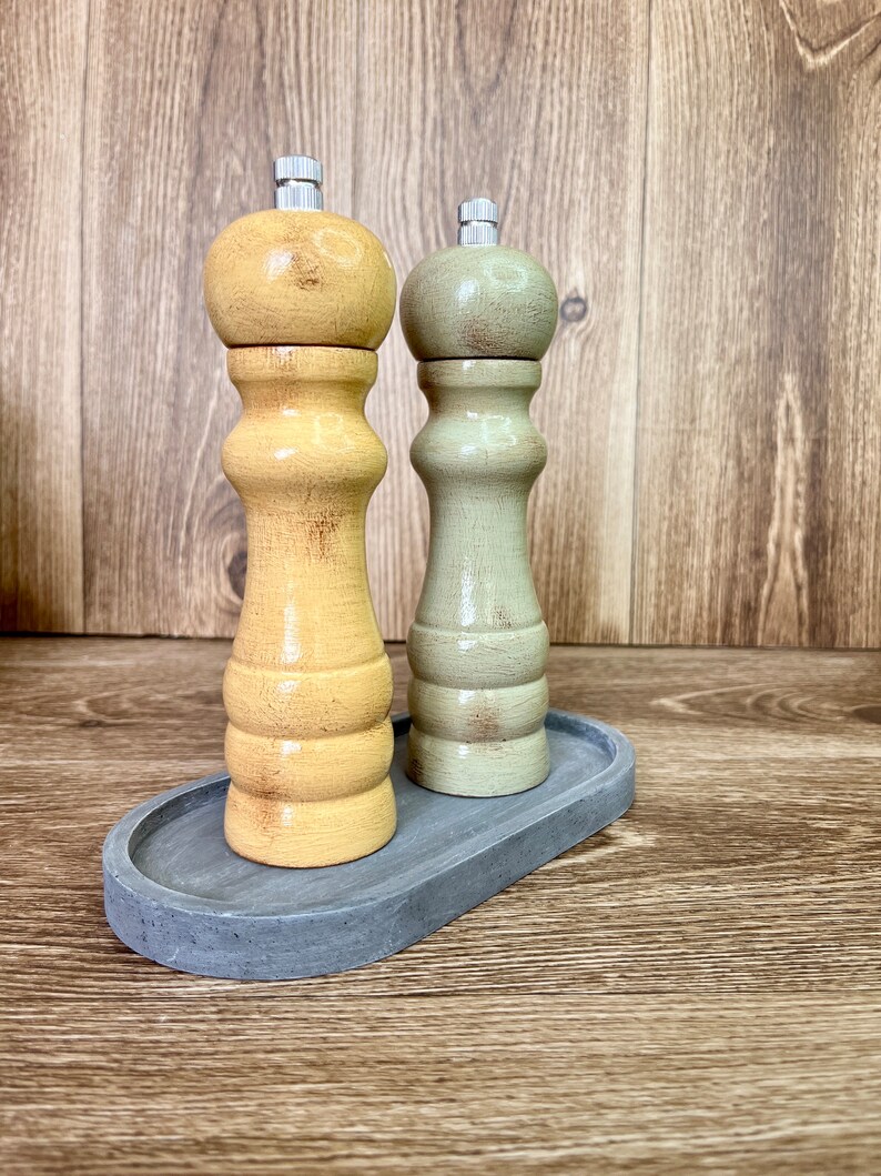 May include: A set of two wooden salt and pepper shakers with a distressed finish. The shakers are in a light yellow and a light green color. The shakers are on a gray oval tray.