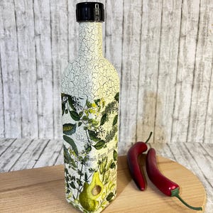 May include: A white ceramic bottle with a black lid and a botanical design. The bottle has a crackled finish and features a green avocado illustration. The bottle is sitting on a wooden cutting board with two red chili peppers.