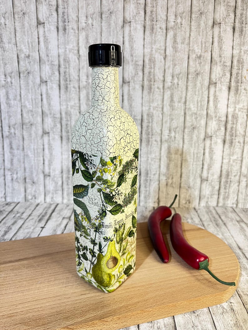 May include: A white ceramic bottle with a black lid and a botanical design. The bottle has a crackled finish and features a green avocado illustration. The bottle is sitting on a wooden cutting board with two red chili peppers.