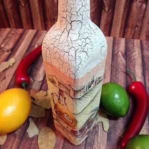May include: A white glass bottle with a cracked paint finish and a green lid. The bottle is decorated with a painted scene of a countryside landscape with a building and trees. The bottle is surrounded by red chilli peppers, green limes, and bay leaves on a wooden surface.