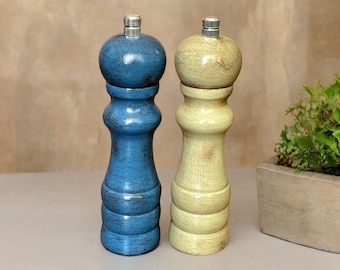 May include: Two wooden salt and pepper grinders with a distressed finish. One grinder is painted in a deep blue color, while the other is a pale yellow. Both feature a rounded top, a contoured body, and a metallic silver adjustment knob.