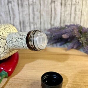 Puede incluir: Una botella de cer&aacute;mica blanca con un dise&ntilde;o negro y marr&oacute;n y un pico de vertido de pl&aacute;stico transparente. La botella est&aacute; sobre una superficie de madera con una tapa de pl&aacute;stico negro al lado.