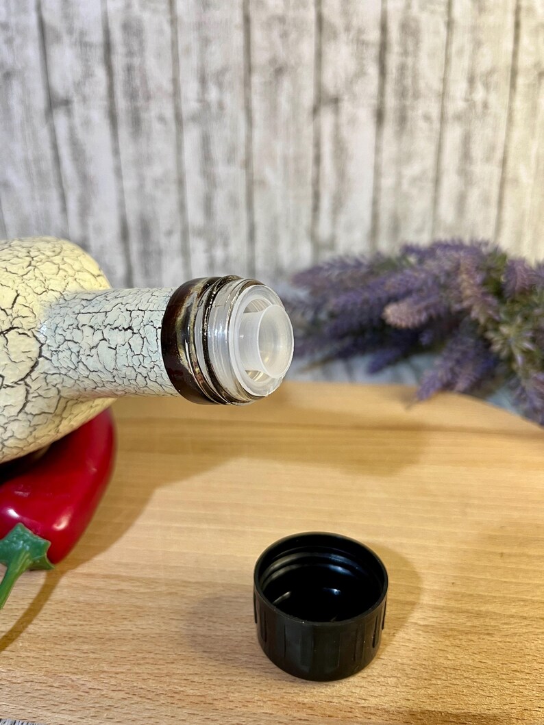 Puede incluir: Una botella de cer&aacute;mica blanca con un dise&ntilde;o negro y marr&oacute;n y un pico de vertido de pl&aacute;stico transparente. La botella est&aacute; sobre una superficie de madera con una tapa de pl&aacute;stico negro al lado.