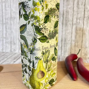 May include: A white glass bottle with a distressed finish and a green and yellow floral design. The bottle has the words "Basil, Lemon, Avocado" printed on it.