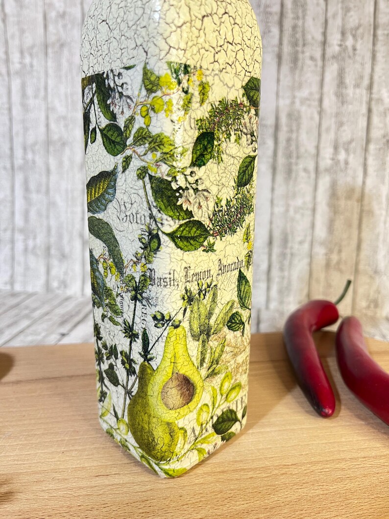 May include: A white glass bottle with a distressed finish and a green and yellow floral design. The bottle has the words "Basil, Lemon, Avocado" printed on it.