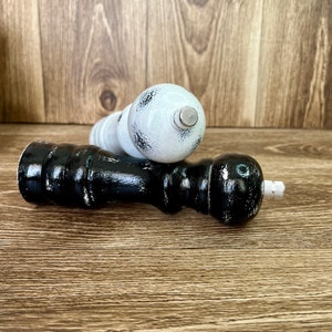 May include: Two wooden pepper mills, one finished in black and the other in white, both featuring a distressed paint effect. Each mill has a rounded top, a contoured body, and a small metallic adjustment knob at the very top end.