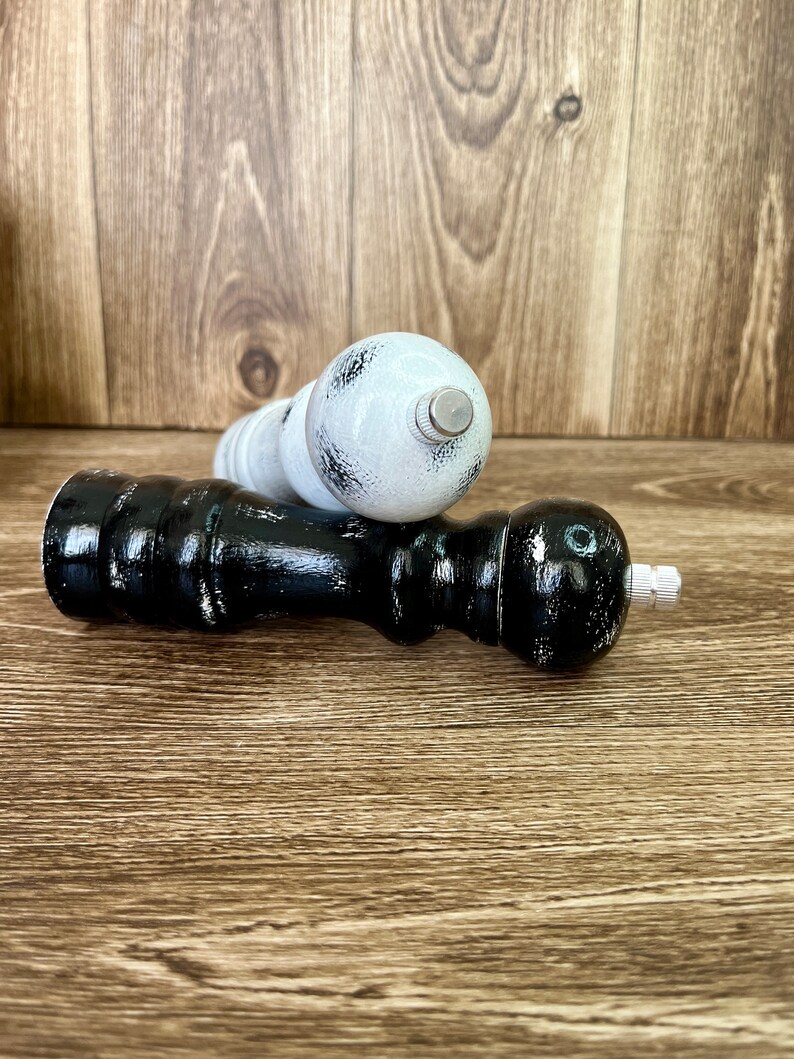 May include: Two wooden pepper mills, one finished in black and the other in white, both featuring a distressed paint effect. Each mill has a rounded top, a contoured body, and a small metallic adjustment knob at the very top end.