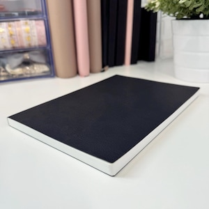 Puede incluir: Un cuaderno negro encuadernado en cuero con bordes blancos. El cuaderno está acostado sobre una superficie blanca.