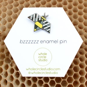 May include: A black and white geometric bee enamel pin with yellow accents. The pin is on a white card with the text "bzzzzzz enamel pin", "whole circle studio", "wholecirclestudio.com", and "@wholecirclestudio".