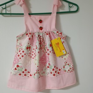 Op de afbeelding: Een roze en witte babyjurk met een aardbeienpatroon. De jurk heeft een gestreept lijfje met twee aardbeivormige knopen en strikbandjes. De rok heeft een patchwork ontwerp met aardbeien en bloemmotieven. Een geel label hangt eraan.