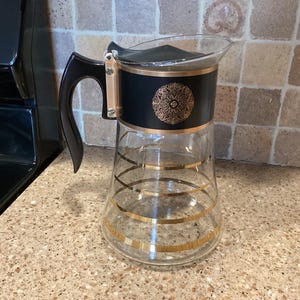 May include: A vintage glass pitcher with a black band featuring a gold medallion design. The pitcher has a black handle and gold stripes. The pitcher is on a speckled countertop.