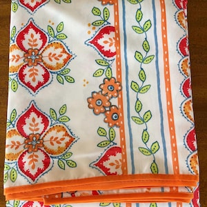 May include: A folded white fabric featuring a repeating pattern of large red and orange floral motifs, alongside vertical stripes of orange bands and green vine designs with small leaves. The fabric is finished with a solid orange border trim.