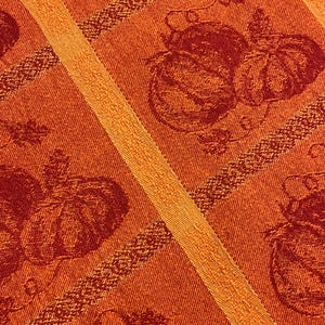 May include: A woven fabric featuring a repeating pattern of pumpkins and vines in shades of deep red and orange. The design is organized into vertical panels separated by textured orange stripes, creating a structured and decorative textile surface.