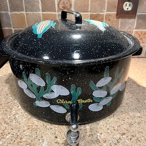 May include: A black speckled enamel stock pot with a matching lid, featuring a spigot at the base. The exterior is decorated with green seaweed and white clam illustrations, with the yellow text Clam Broth printed on the front side.