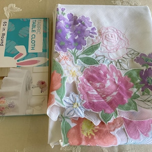 May include: A white fabric table cloth with embroidered floral designs in pink, purple, and green. The text "FABRIC TABLE CLOTH" and "60 in. Round" is printed on the packaging.