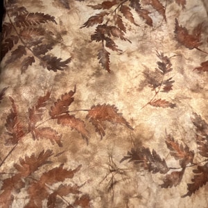 May include: A piece of fabric featuring a mottled, light tan and beige background with a repeating pattern of dark brown fern leaves. The design displays a textured, organic appearance with varying shades of brown creating depth across the surface.