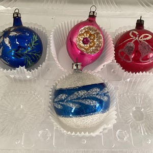 May include: A collection of vintage Christmas ornaments in a clear plastic container. The ornaments are in various shapes and colors, including blue, pink, and red. Each ornament has a unique design, such as painted leaves, a floral pattern, and bells.