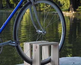 May include: A blue road bicycle with silver components and black tires is displayed. The front wheel rests in a light-colored wooden bike stand. The background features a lake and trees.