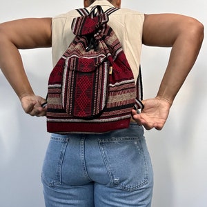 Puede incluir: Una mochila tejida de color rojo, negro y beige con cierre de cordón. La mochila tiene un pequeño bolsillo en la parte delantera.