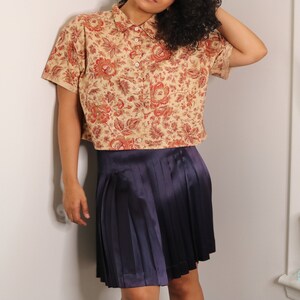 May include: A cropped floral button-down shirt with a beige background and red and orange flowers. The shirt is paired with a dark purple pleated skirt.