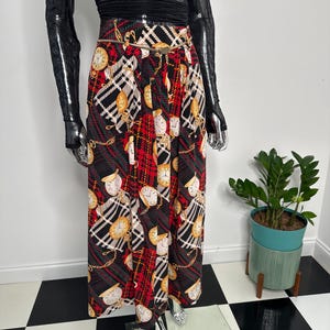 May include: A long, pleated skirt with a black, red, and white plaid pattern. The skirt features a repeating design of gold-colored pocket watches and gold chain accents. A gold-colored belt cinches the waist. The skirt is part of a vintage clothing collection.