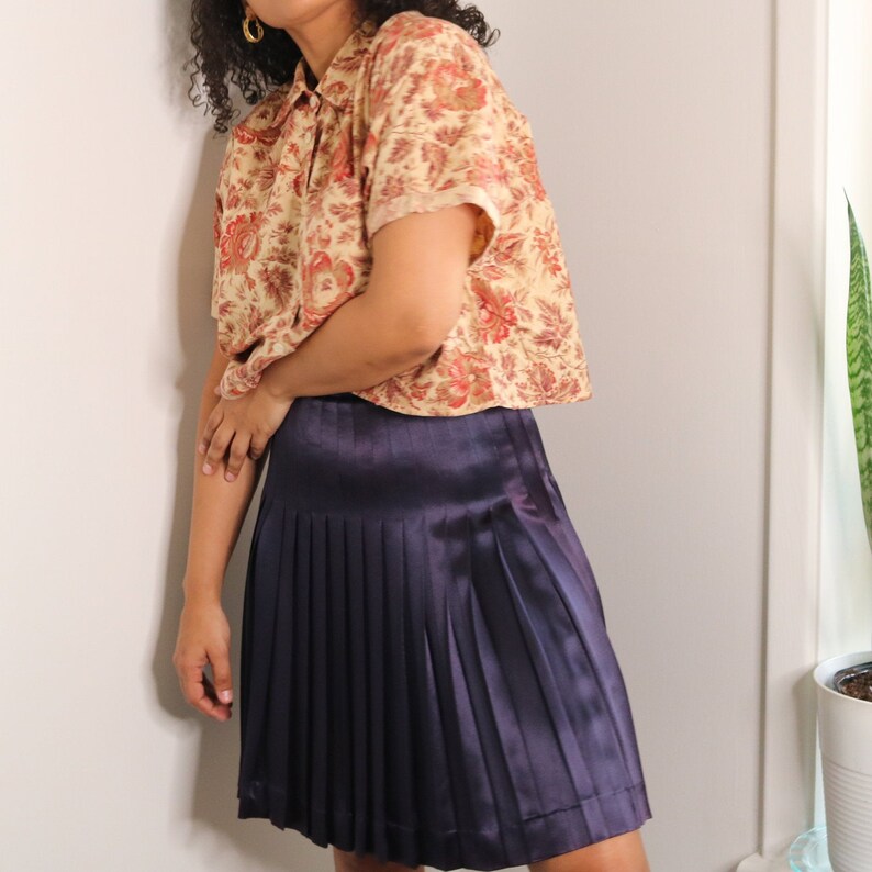 May include: A woman wearing a short-sleeved, floral print button-down shirt and a dark purple pleated skirt.