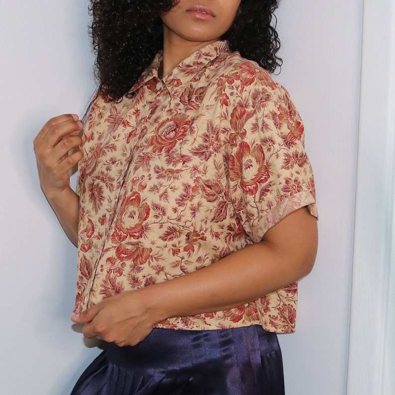 May include: A cropped, short-sleeved button-down shirt with a floral print in shades of red, brown, and cream. The shirt has a relaxed fit and a collar.