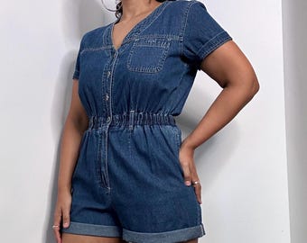 May include: A blue denim jumpsuit with short sleeves, a button-up front, and a cinched waist. The jumpsuit has a pocket on the left chest and rolled-up cuffs.