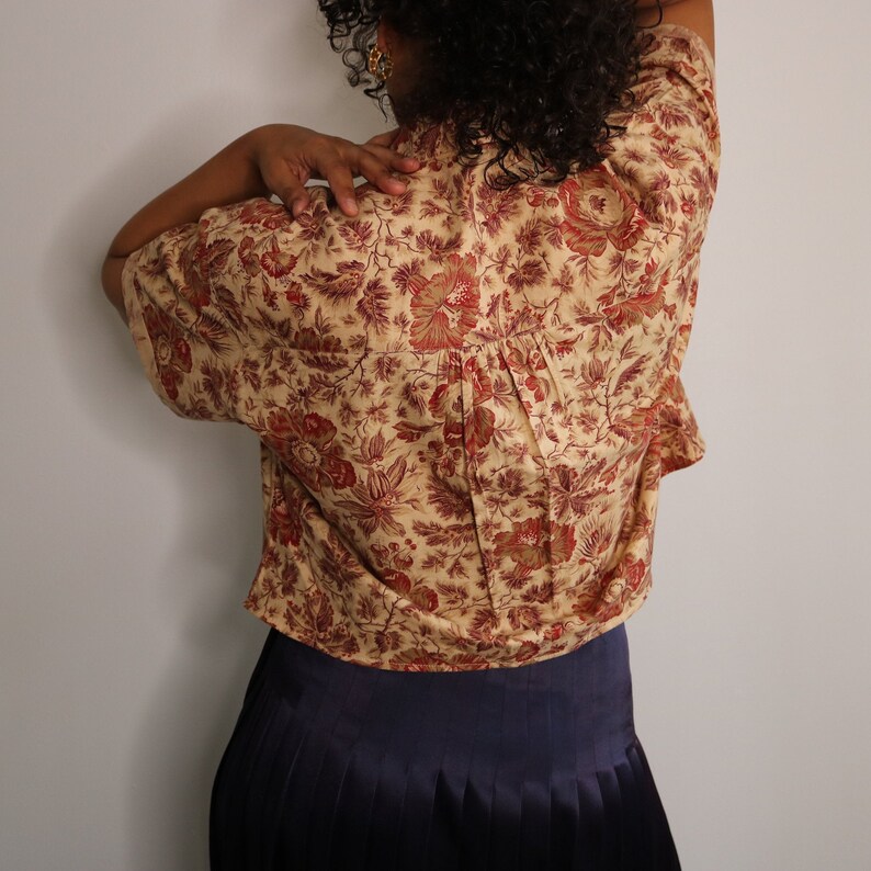 May include: A cropped floral print shirt with a gathered back detail. The shirt is beige with a red and brown floral pattern. The model is wearing a dark blue pleated skirt.